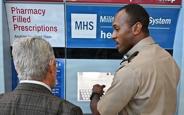Two individuals are standing in front of an automated pharmacy prescription pickup kiosk branded with MHS (Military Health System) signage. One person in a tan/khaki uniform appears to be demonstrating or explaining the kiosk's operation to another person in a gray suit jacket. The setting appears to be a military health or expo event, with additional banners visible in the background.
