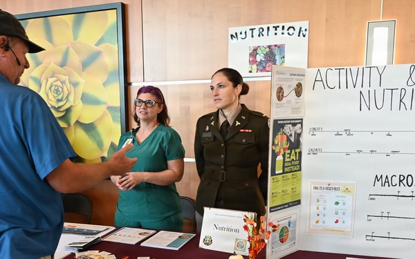 A health fair booth operated by Army Public Health Nursing from Fort Cavazos, Texas, where two women — one in military uniform and one in green scrubs — are speaking with a civilian visitor. The booth features educational displays on Activity &amp; Nutrition, Macros, and dietary information. The table is covered with a maroon tablecloth branded with the Army Public Health Nursing logo and is decorated with fall pumpkins and informational pamphlets.