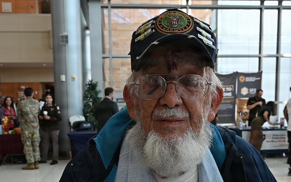 An elderly man wearing a U.S. Army veteran's cap and glasses stands in the foreground of what appears to be a veterans' event or resource fair held in a modern building lobby. He has a white beard and appears to have a small wound or mark on his forehead. In the background, groups of people including individuals in military uniforms are gathered near informational tables and displays.