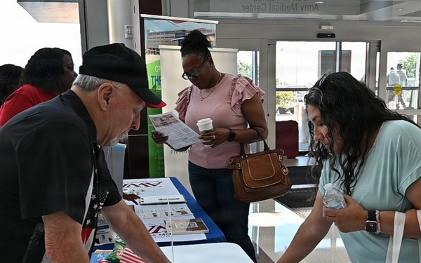 An indoor health fair or informational event is taking place in what appears to be a medical center lobby, visible from signage reading 'Army Medical Center' in the background. A man in a black shirt and hat is presenting materials at a table covered with a white tablecloth to a woman in a teal shirt carrying a white tote bag. A second woman in a pink top stands nearby reviewing a pamphlet while holding a coffee cup.