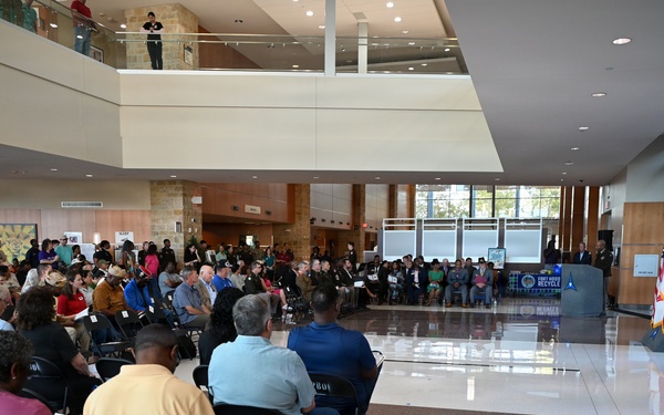 A large indoor gathering is taking place in a modern building lobby or atrium, with a crowd of attendees seated in folding chairs facing a speaker at a podium. A military flag and banner are visible near the podium, suggesting this is an official or ceremonial event. The venue features a two-story open atrium with observers on the upper level balcony.