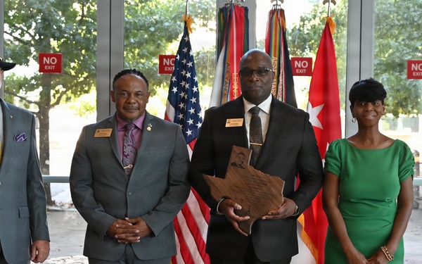 Four individuals are posed for a formal group photo in what appears to be a government or military building lobby. One person in the center is holding a wooden award shaped like the state of Texas. The setting includes American and military flags in the background, suggesting an official recognition or awards ceremony.