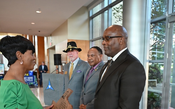 A formal ceremony is taking place in what appears to be a government or military building lobby. A woman in a green dress is presenting or receiving a wooden plaque/award to/from a man in a dark suit, while two other men stand nearby as witnesses. An American flag and what appears to be a military unit flag are visible in the background.