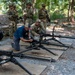 Military personnel in camouflage uniforms are engaged in a weapons training exercise at an outdoor range with a gravel and wooded setting. Several soldiers are kneeling and working on tripod-mounted heavy machine guns laid out on wooden boards. A civilian instructor in a blue shirt and cap appears to be supervising or demonstrating proper weapon handling techniques.