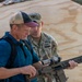 A U.S. Army soldier in OCP (Operational Camouflage Pattern) uniform is showing or demonstrating a modern semi-automatic rifle to a civilian man wearing a blue polo shirt and baseball cap. The two individuals are examining the firearm together under what appears to be a temporary shelter or tent structure with plywood backing. The setting suggests a military demonstration, equipment showcase, or familiarization event.