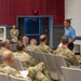 A civilian presenter in a blue polo shirt addresses a group of military personnel seated in an auditorium or multipurpose hall. The audience consists predominantly of soldiers in U.S. Army OCP (Operational Camouflage Pattern) uniforms, suggesting a briefing, training session, or informational meeting. The setting appears to be a military installation facility with a stage area visible to the right.