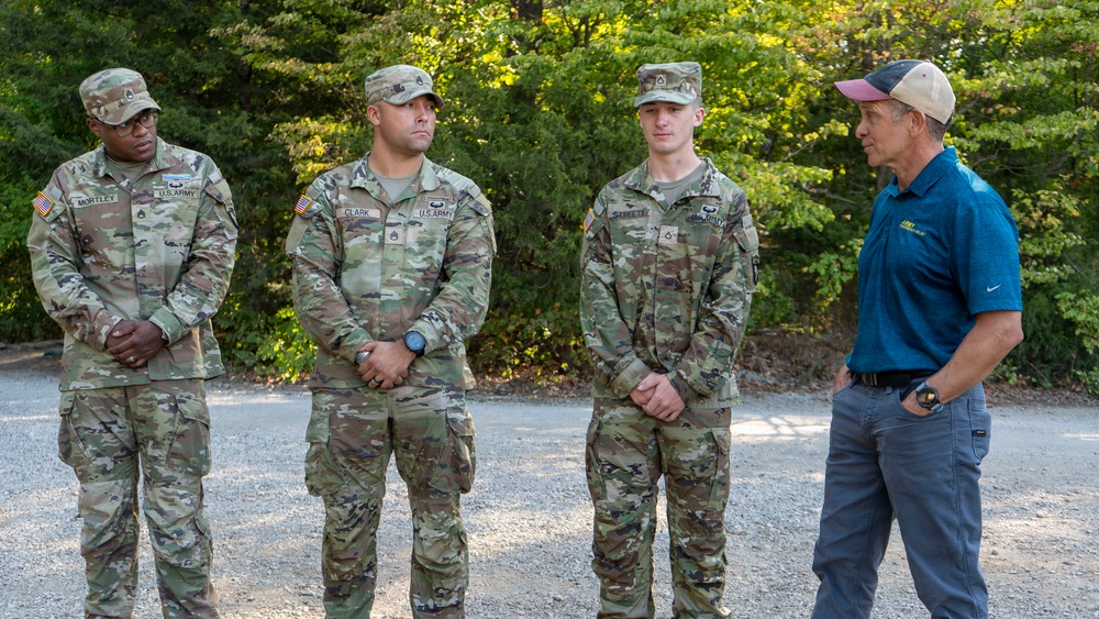 Three U.S. Army soldiers in OCP (Operational Camouflage Pattern) uniforms are standing at attention while a civilian man in a blue polo shirt and baseball cap addresses them outdoors. The soldiers' name tapes are partially visible, reading 'Mortley,' 'Clark,' and 'Street.' The setting appears to be a gravel area surrounded by trees, suggesting a training or briefing environment.