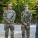 Three U.S. Army soldiers in OCP (Operational Camouflage Pattern) uniforms are standing at attention while a civilian man in a blue polo shirt and baseball cap addresses them outdoors. The soldiers' name tapes are partially visible, reading 'Mortley,' 'Clark,' and 'Street.' The setting appears to be a gravel area surrounded by trees, suggesting a training or briefing environment.