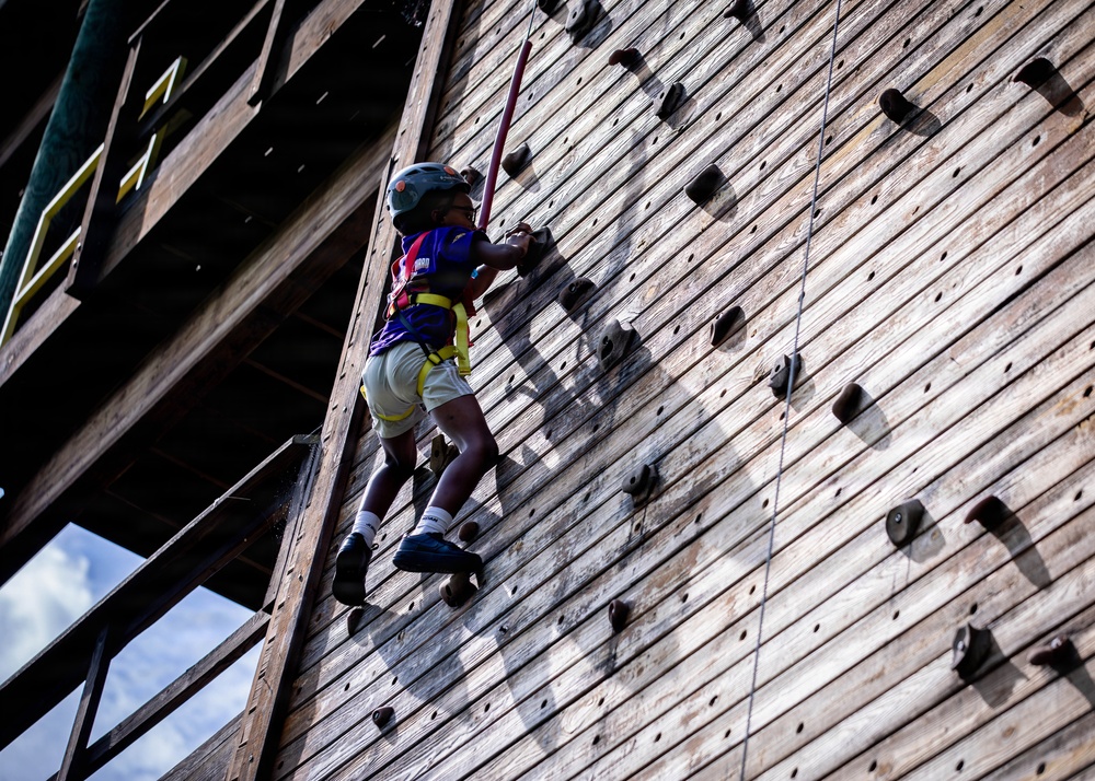 A child is climbing a tall wooden climbing wall equipped with hand and foot holds, secured by a safety harness and wearing a helmet. The photo is taken from a low angle looking upward, emphasizing the height of the structure. The setting appears to be an outdoor adventure or summer camp facility.