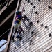 A child is climbing a tall wooden climbing wall equipped with hand and foot holds, secured by a safety harness and wearing a helmet. The photo is taken from a low angle looking upward, emphasizing the height of the structure. The setting appears to be an outdoor adventure or summer camp facility.