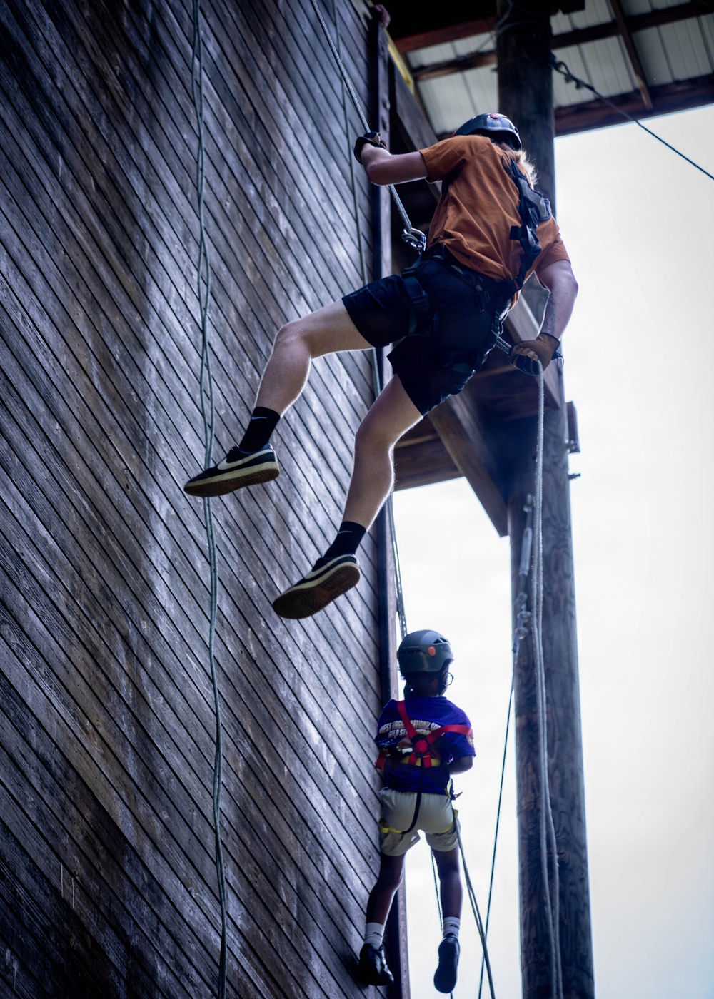 Two individuals are rappelling or climbing down the exterior wooden wall of a tall building, both equipped with safety harnesses and helmets. The upper climber wears an orange shirt and dark shorts, while the lower climber appears smaller in stature and wears a purple shirt with a colorful harness. The image is shot from a low angle, emphasizing the height and vertical scale of the activity.