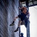 Two individuals are rappelling or climbing down the exterior wooden wall of a tall building, both equipped with safety harnesses and helmets. The upper climber wears an orange shirt and dark shorts, while the lower climber appears smaller in stature and wears a purple shirt with a colorful harness. The image is shot from a low angle, emphasizing the height and vertical scale of the activity.