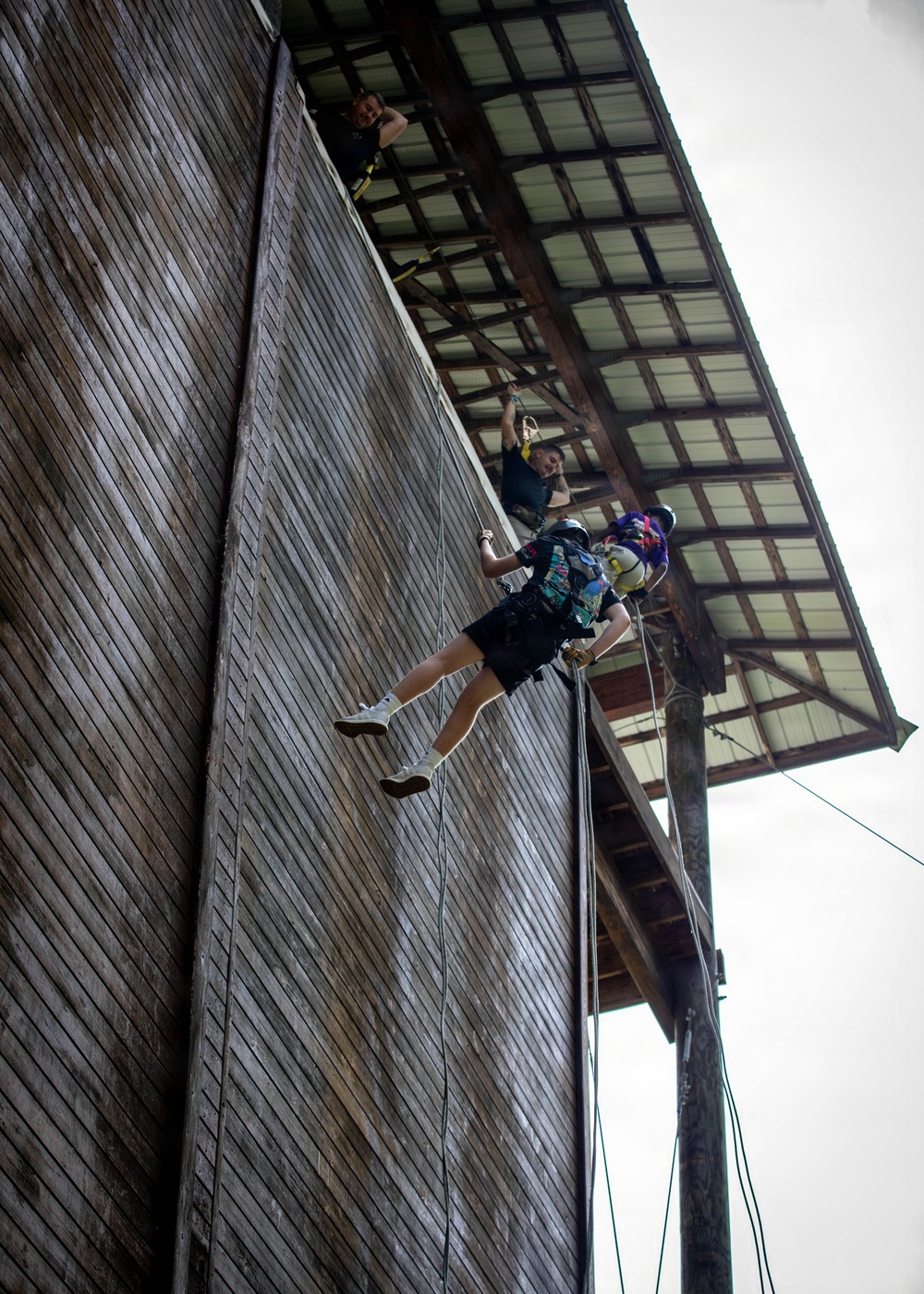 A person is rappelling down the exterior wooden wall of a large barn-like structure, suspended by ropes and wearing a harness, helmet, and colorful backpack. At least two other individuals are visible at the top edge of the building, appearing to assist or observe the descent. The activity appears to be a recreational or camp-based rappelling exercise.