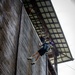 A person is rappelling down the exterior wooden wall of a large barn-like structure, suspended by ropes and wearing a harness, helmet, and colorful backpack. At least two other individuals are visible at the top edge of the building, appearing to assist or observe the descent. The activity appears to be a recreational or camp-based rappelling exercise.