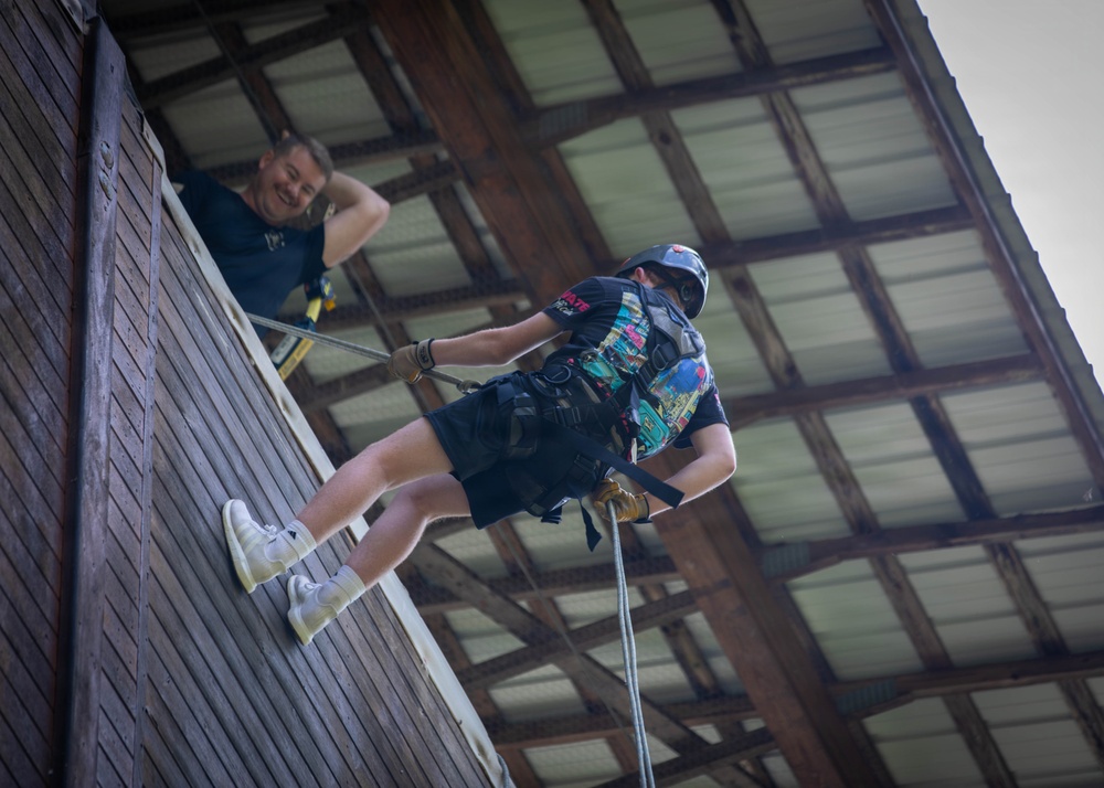 A young person is rappelling down a wooden wall inside a covered structure, wearing a helmet, harness, gloves, and casual clothing including white sneakers. An adult instructor or supervisor leans over the top edge of the wall, smiling and observing the descent. The setting appears to be an indoor or covered training/recreational rappelling tower.