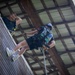 A young person is rappelling down a wooden wall inside a covered structure, wearing a helmet, harness, gloves, and casual clothing including white sneakers. An adult instructor or supervisor leans over the top edge of the wall, smiling and observing the descent. The setting appears to be an indoor or covered training/recreational rappelling tower.