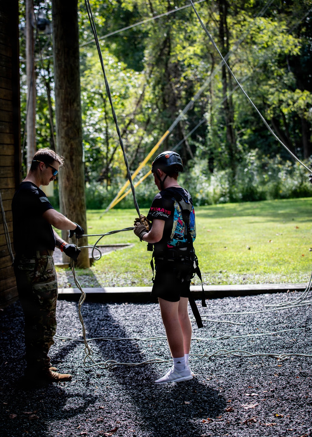 Two individuals are engaged in a ropes course or zip-line activity in an outdoor wooded setting. One person wearing camouflage pants and gloves appears to be an instructor managing ropes, while the other person is equipped with a safety harness, helmet, and gloves as a participant. Multiple cables and ropes extend upward and across the scene, indicating an aerial adventure course setup.