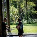 Two individuals are engaged in a ropes course or zip-line activity in an outdoor wooded setting. One person wearing camouflage pants and gloves appears to be an instructor managing ropes, while the other person is equipped with a safety harness, helmet, and gloves as a participant. Multiple cables and ropes extend upward and across the scene, indicating an aerial adventure course setup.