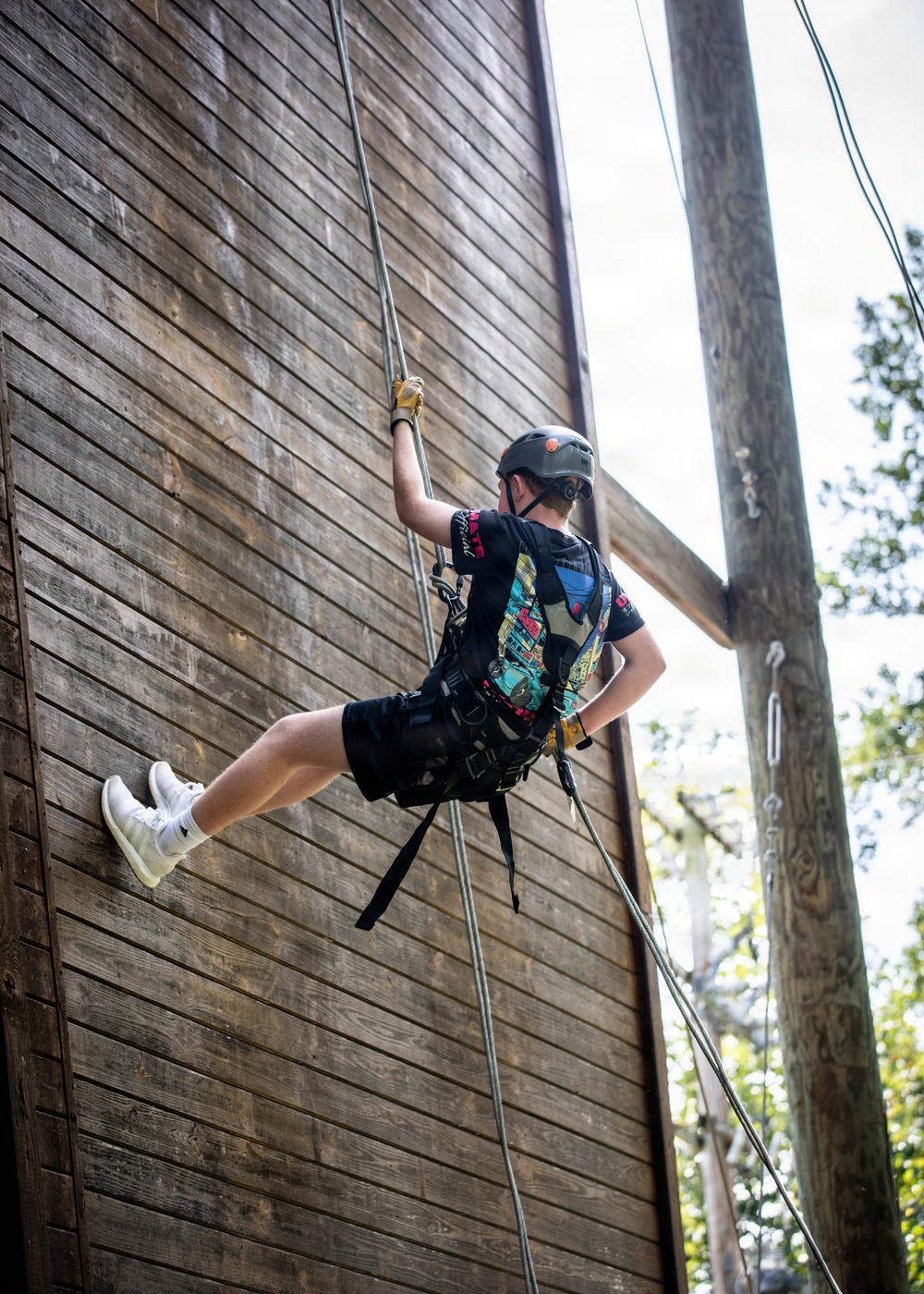 A young person is rappelling or climbing down a large wooden wall structure using ropes and a safety harness. They are equipped with standard climbing safety gear and appear to be engaged in a recreational or camp-based outdoor activity. One foot is braced against the wooden wall while the other is extended outward.
