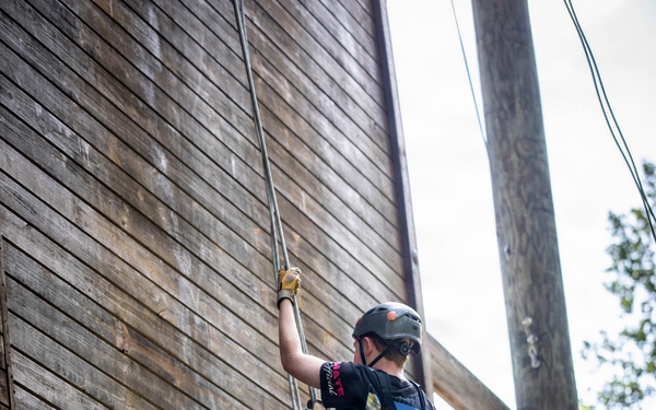 A young person is rappelling or climbing down a large wooden wall structure using ropes and a safety harness. They are equipped with standard climbing safety gear and appear to be engaged in a recreational or camp-based outdoor activity. One foot is braced against the wooden wall while the other is extended outward.
