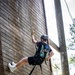A young person is rappelling or climbing down a large wooden wall structure using ropes and a safety harness. They are equipped with standard climbing safety gear and appear to be engaged in a recreational or camp-based outdoor activity. One foot is braced against the wooden wall while the other is extended outward.