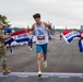 A male runner wearing bib number 5493 and a 'Dayton' singlet crosses the finish line of the U.S. Air Force Marathon, breaking through a red, white, and blue finisher's tape. He is gesturing with both index fingers pointed upward in a celebratory manner, suggesting he may be the first-place finisher. A uniformed U.S. Air Force soldier on the left and a race official on the right hold the finish tape.
