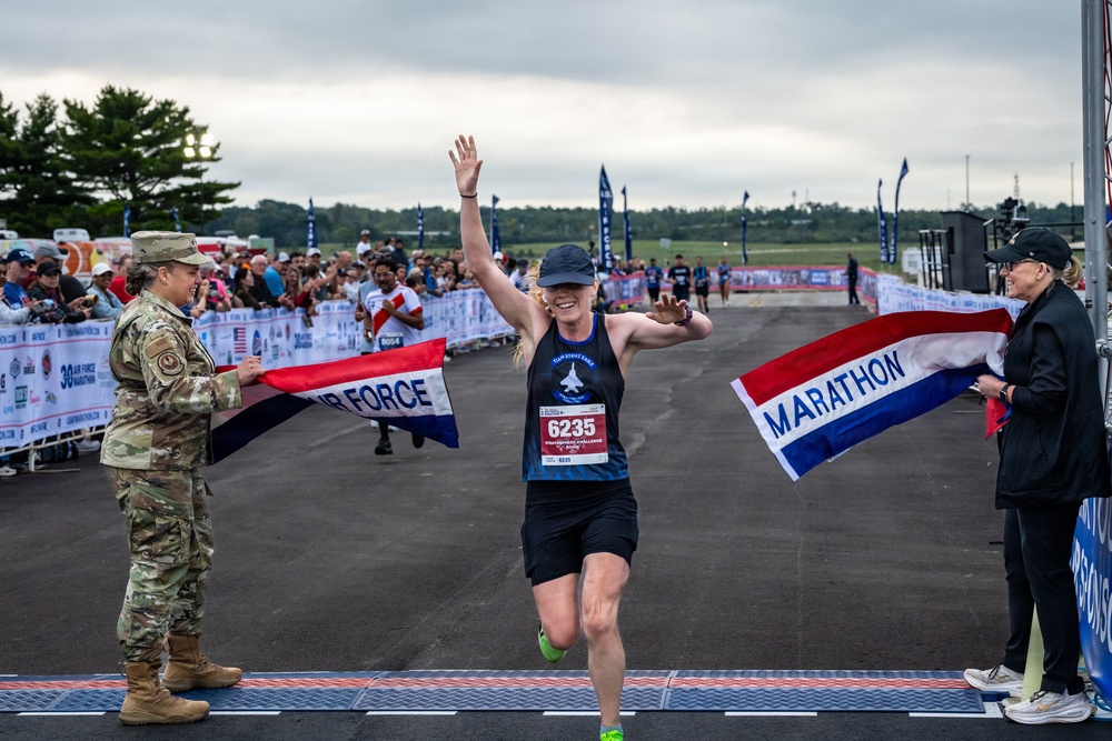 A female runner wearing bib number 6235 crosses the finish line of the Air Force Marathon, arms raised in celebration. She is breaking through a red, white, and blue ribbon held by a military service member in camouflage uniform on the left and a civilian on the right. A large crowd of spectators lines the course in the background.