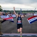 A female runner wearing bib number 6235 crosses the finish line of the Air Force Marathon, arms raised in celebration. She is breaking through a red, white, and blue ribbon held by a military service member in camouflage uniform on the left and a civilian on the right. A large crowd of spectators lines the course in the background.
