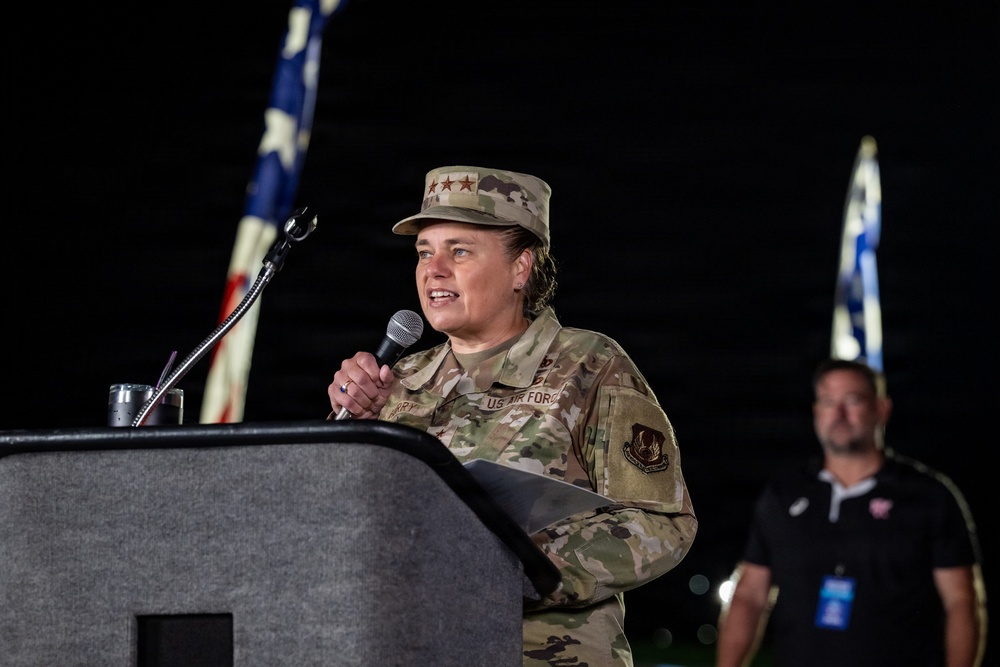 A U.S. Air Force officer in OCP (Operational Camouflage Pattern) uniform is speaking at a podium while holding a microphone. The officer's cap displays what appears to be three stars, indicating a high-ranking general officer. The setting appears to be an outdoor nighttime event with American and other flags visible in the background.