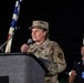 A U.S. Air Force officer in OCP (Operational Camouflage Pattern) uniform is speaking at a podium while holding a microphone. The officer's cap displays what appears to be three stars, indicating a high-ranking general officer. The setting appears to be an outdoor nighttime event with American and other flags visible in the background.