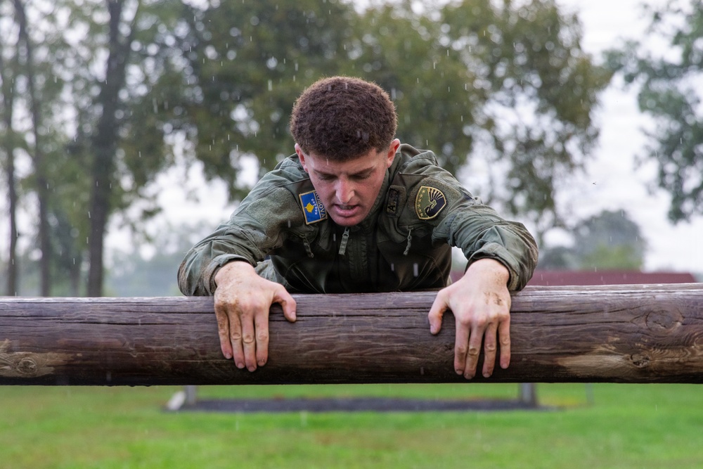 A young male soldier in olive/green military uniform is navigating an outdoor obstacle course in rainy conditions, pulling himself over a large wooden log barrier. His uniform displays at least two visible patches, including what appears to be a flag patch on the left shoulder. The image captures a moment of physical exertion and determination during what appears to be military training.