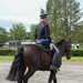 A uniformed rider, appearing to be a military or ceremonial officer, is mounted on a dark brown/black horse in an outdoor setting. The rider is viewed from behind, wearing a formal black jacket, blue trousers with a gold stripe, tall black riding boots, and a peaked cap. The scene appears to take place at a formal equestrian or ceremonial venue with green trees and manicured grounds in the background.