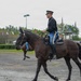 A military rider in formal dress uniform is mounted on a dark brown horse, moving across a gravel arena or parade ground. The rider wears a black jacket, blue trousers with a yellow stripe, black boots, and a peaked cap consistent with U.S. Army ceremonial dress. A second horse and rider are partially visible in the background, suggesting a formal equestrian event or military demonstration.