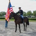 A uniformed rider on a dark brown horse holds the reins during what appears to be a ceremonial event. A person standing on the ground holds an American flag with gold fringe on a pole beside the horse. The setting appears to be a gravel area at what may be a fairground or equestrian venue.