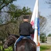 A military soldier in dress uniform is mounted on a dark bay horse, carrying a large ceremonial flag with gold fringe. The flag appears to be a U.S. Army unit colors or organizational flag, partially visible with text reading 'WE'LL DEFEND' and featuring blue and red insignia. The scene is photographed from behind along a gravel path surrounded by trees and grass.