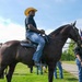 A man wearing a yellow cowboy hat, black t-shirt, and jeans is seated on a large dark brown horse in an outdoor grassy setting. Several other people are visible nearby, including one person holding an American flag. The scene appears to be an organized outdoor equestrian or community event.