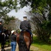 Two uniformed riders on horseback are positioned in a tree-lined area, accompanied by a civilian woman wearing a white cowboy hat and jeans. An American flag is visible between the horses, suggesting a ceremonial or official event. The setting appears to be an outdoor venue with manicured grass and mature trees.