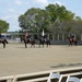 A formal military color guard ceremony is taking place on a polo ground, with four uniformed riders on dark horses carrying flags including the American flag. The event appears to be a pre-match ceremony at an equestrian polo venue, with spectators visible in the foreground and sponsor signage along the arena fence. The setting is an outdoor sand arena on a partly cloudy day with lush green trees in the background.