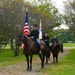 Three uniformed riders on dark horses proceed along a gravel path through a tree-lined grounds, carrying ceremonial flags including the United States flag and what appears to be a military or organizational guidon. The riders wear matching dark dress uniforms with peaked caps, suggesting a formal color guard or ceremonial procession. The setting appears to be a well-maintained estate or military installation with manicured lawns.