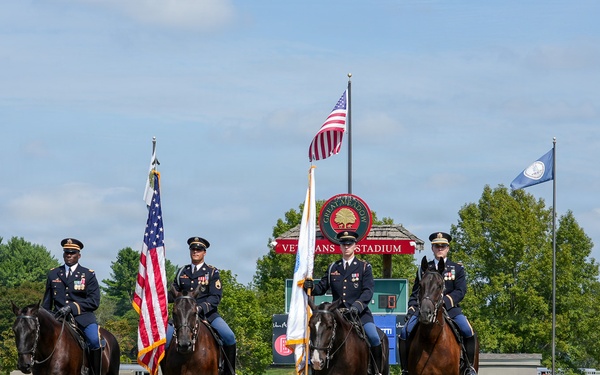Four military personnel in dress uniforms are mounted on dark bay horses in a sandy arena, performing a color guard ceremony. Two riders carry flags — the American flag and what appears to be a military unit flag. The event is taking place at Great Meadow Veterans Stadium, with sponsor banners and flags visible in the background.