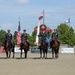 Four military personnel in dress uniforms are mounted on dark bay horses in a sandy arena, performing a color guard ceremony. Two riders carry flags — the American flag and what appears to be a military unit flag. The event is taking place at Great Meadow Veterans Stadium, with sponsor banners and flags visible in the background.