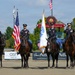 Four military personnel in dress uniforms are mounted on dark horses at what appears to be Great Meadow Veterans Stadium in The Plains, Virginia. Two soldiers are saluting while two others carry flags, including the American flag and what appears to be a military unit flag. The scene depicts a formal military color guard ceremony at an equestrian or sporting venue.