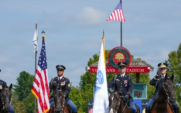 Four U.S. Army soldiers in dress uniforms are mounted on dark horses in a formal color guard ceremony at Great Meadow Veterans Stadium. Two soldiers are saluting while two carry flags, including the American flag and what appears to be a military or organizational flag. The event takes place in an equestrian arena with sponsor banners visible in the background.