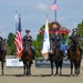 Four U.S. Army soldiers in dress uniforms are mounted on dark horses in a formal color guard ceremony at Great Meadow Veterans Stadium. Two soldiers are saluting while two carry flags, including the American flag and what appears to be a military or organizational flag. The event takes place in an equestrian arena with sponsor banners visible in the background.