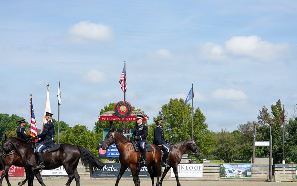 A military color guard ceremony is taking place at an outdoor equestrian arena identified as 'Veterans Stadium.' Four uniformed military riders on dark horses are performing a mounted color guard presentation, carrying flags including the American flag. The event appears to be a formal ceremonial display at what looks like a horse show or equestrian competition venue.