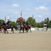 A military color guard ceremony is taking place at an outdoor equestrian arena identified as 'Veterans Stadium.' Four uniformed military riders on dark horses are performing a mounted color guard presentation, carrying flags including the American flag. The event appears to be a formal ceremonial display at what looks like a horse show or equestrian competition venue.