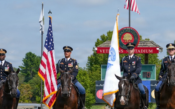 Four military personnel in dress uniforms are mounted on dark bay horses in a formal color guard ceremony at Great Meadow Events venue. Two riders carry flags — the United States flag and what appears to be a military or organizational flag. The event takes place in an equestrian arena surrounded by sponsor banners and a stadium sign reading 'Veterans Stadium'.