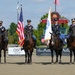 Four military personnel in dress uniforms are mounted on dark bay horses in a formal color guard ceremony at Great Meadow Events venue. Two riders carry flags — the United States flag and what appears to be a military or organizational flag. The event takes place in an equestrian arena surrounded by sponsor banners and a stadium sign reading 'Veterans Stadium'.