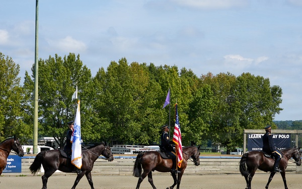 A formal military or ceremonial color guard on horseback is performing in an outdoor equestrian arena. Four riders on dark horses are parading in formation, with at least two carrying flags including a visible American flag. The setting appears to be a polo or equestrian facility based on visible signage.