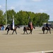A formal military or ceremonial color guard on horseback is performing in an outdoor equestrian arena. Four riders on dark horses are parading in formation, with at least two carrying flags including a visible American flag. The setting appears to be a polo or equestrian facility based on visible signage.
